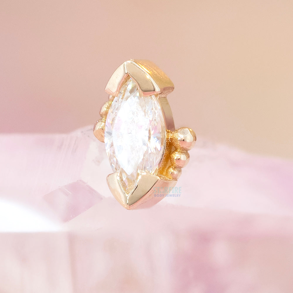 A threadless yellow gold end featuring a marquise-cut white cz gemstone in a decorative setting, resting on a soft pink surface against a matching pink background.