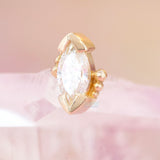 A threadless yellow gold end featuring a marquise-cut white cz gemstone in a decorative setting, resting on a soft pink surface against a matching pink background.